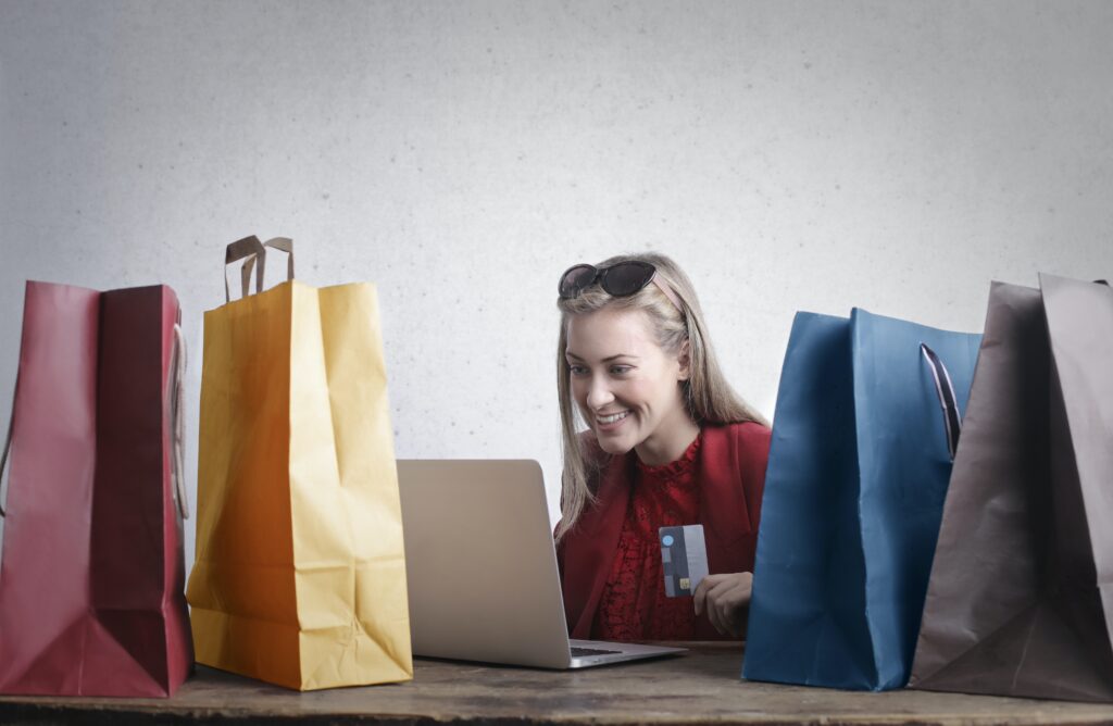online vs high street shopping - woman sitting at laptop with a credit card surrounded by paper shopping bags