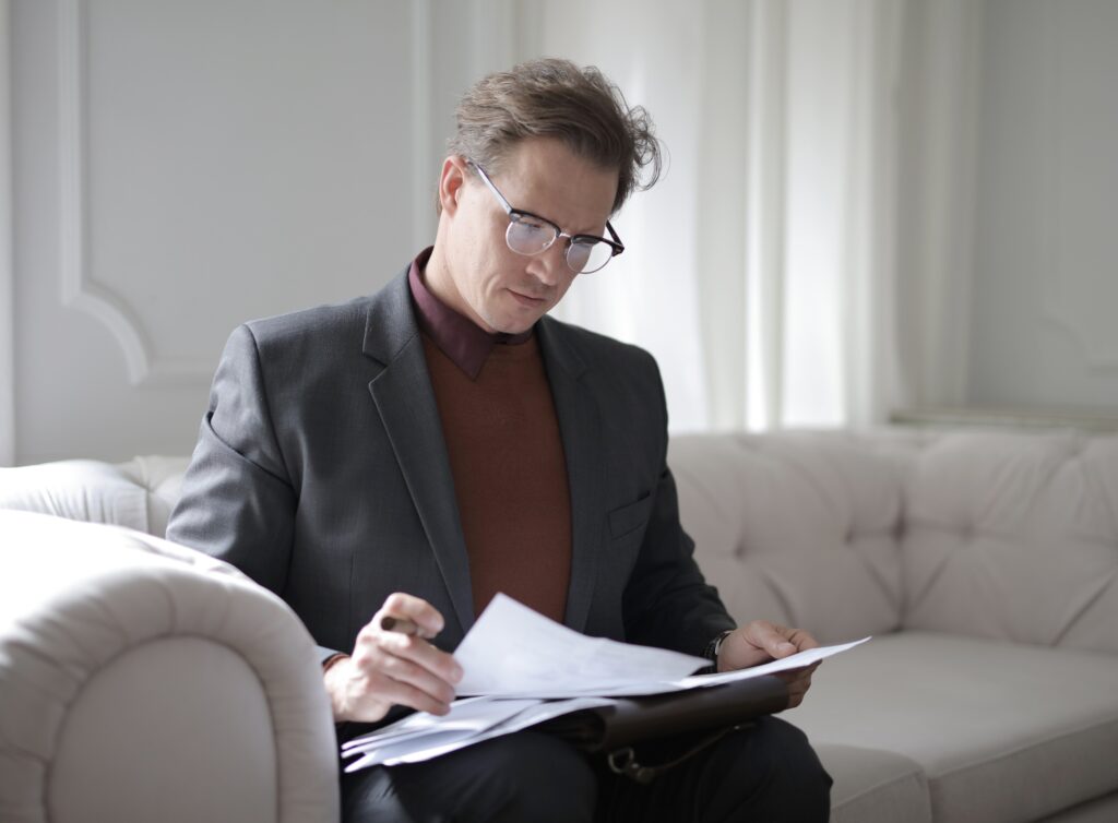 Most efficient way to pay yourself - man in a suit and polo neck with glasses sitting on a beige sofa in a minimalist room reading paperwork