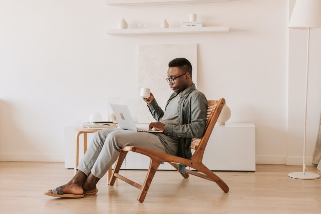 Bookkeep for Xero - Man sitting in a wooden chair in the centre of a minimalist room, working on a laptop with a cup of coffee in his hand