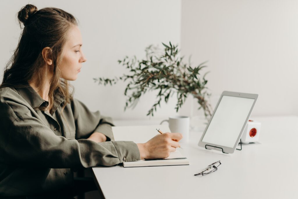 VAT rules for e-commerce - lady sitting at a minimalist desk with a coffee, notebook, tablet propped up to look at the screen and a pot plant in the background
