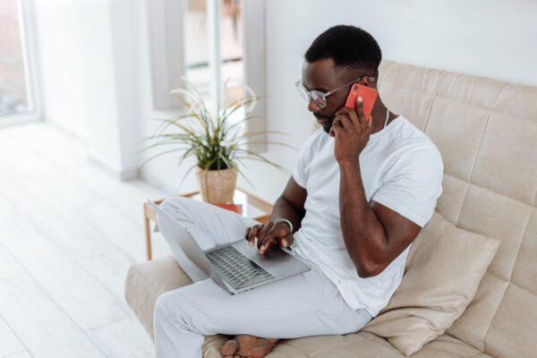 Deferred VAT payment for e-commerce - man dressed in white sitting on a beige armchair in a minimalist room on the phone with a laptop on his lap
