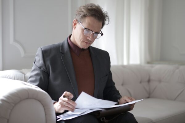 Most efficient way to pay yourself - man in a suit and polo neck with glasses sitting on a beige sofa in a minimalist room reading paperwork