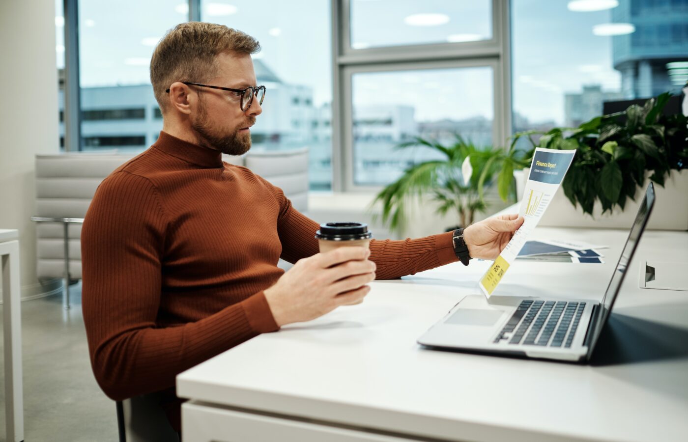 Top Xero integrations for e-commerce - man in a brown tutleneck sitting at a desk with a coffee and a laptop in a spacious office with a huge window