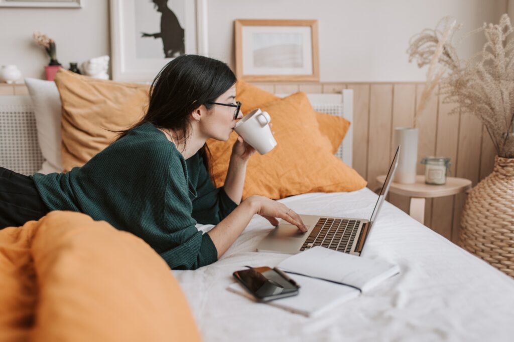 Bookkeep for Xero - young woman sipping coffee in one hand lying on a bed with white duvet and orange throw pillows working on a laptop with a notebook beside her