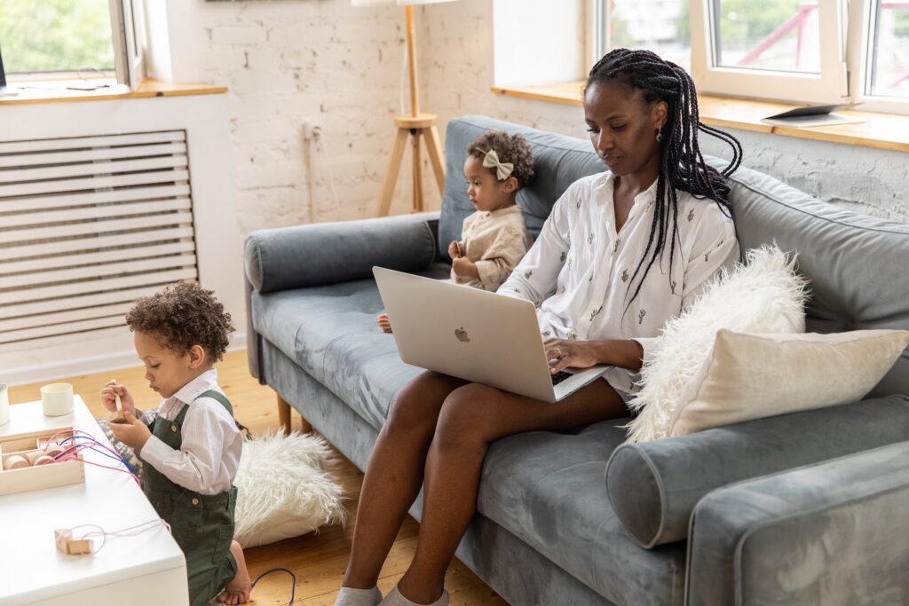 What are e-commerce accountants - black woman in a white shite with long hair sitting on a sofa, laptop on her knee and two young children around her