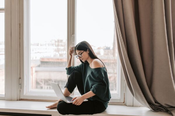 Does Helium 10 work in the UK - woman in black off the shoulder jumper, dark hair and glasses sitting in a window space working on a laptop and thinking