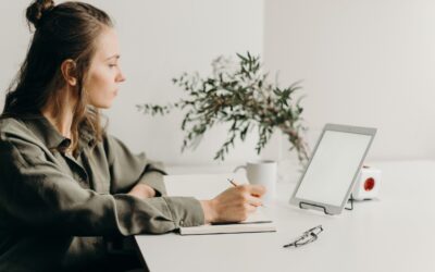 VAT rules for e-commerce - lady sitting at a minimalist desk with a coffee, notebook, tablet propped up to look at the screen and a pot plant in the background