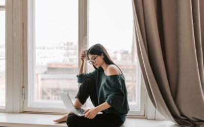 Does Helium 10 work in the UK - woman in black off the shoulder jumper, dark hair and glasses sitting in a window space working on a laptop and thinking